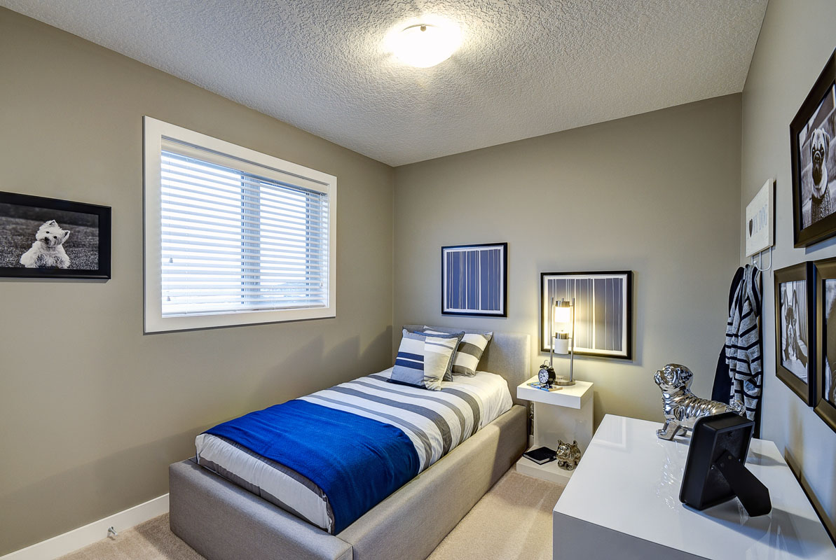 Bedroom 1 with a made twinbed with a blue comforter and a lit lamp on a nightstand next to the bed. Banbury ll model home from Nuvista Homes.