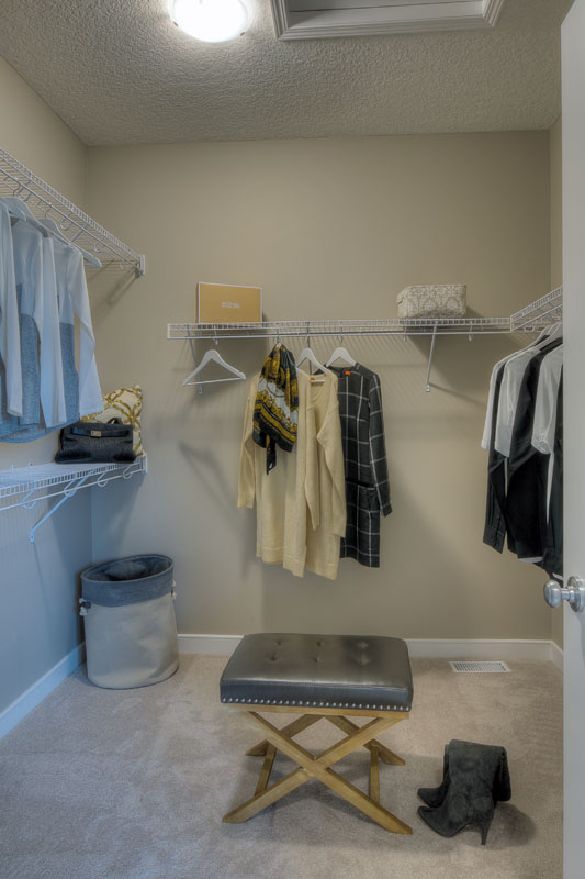 Walk in closet in the Bentley II model home with small leather stool and clothing hanging up.