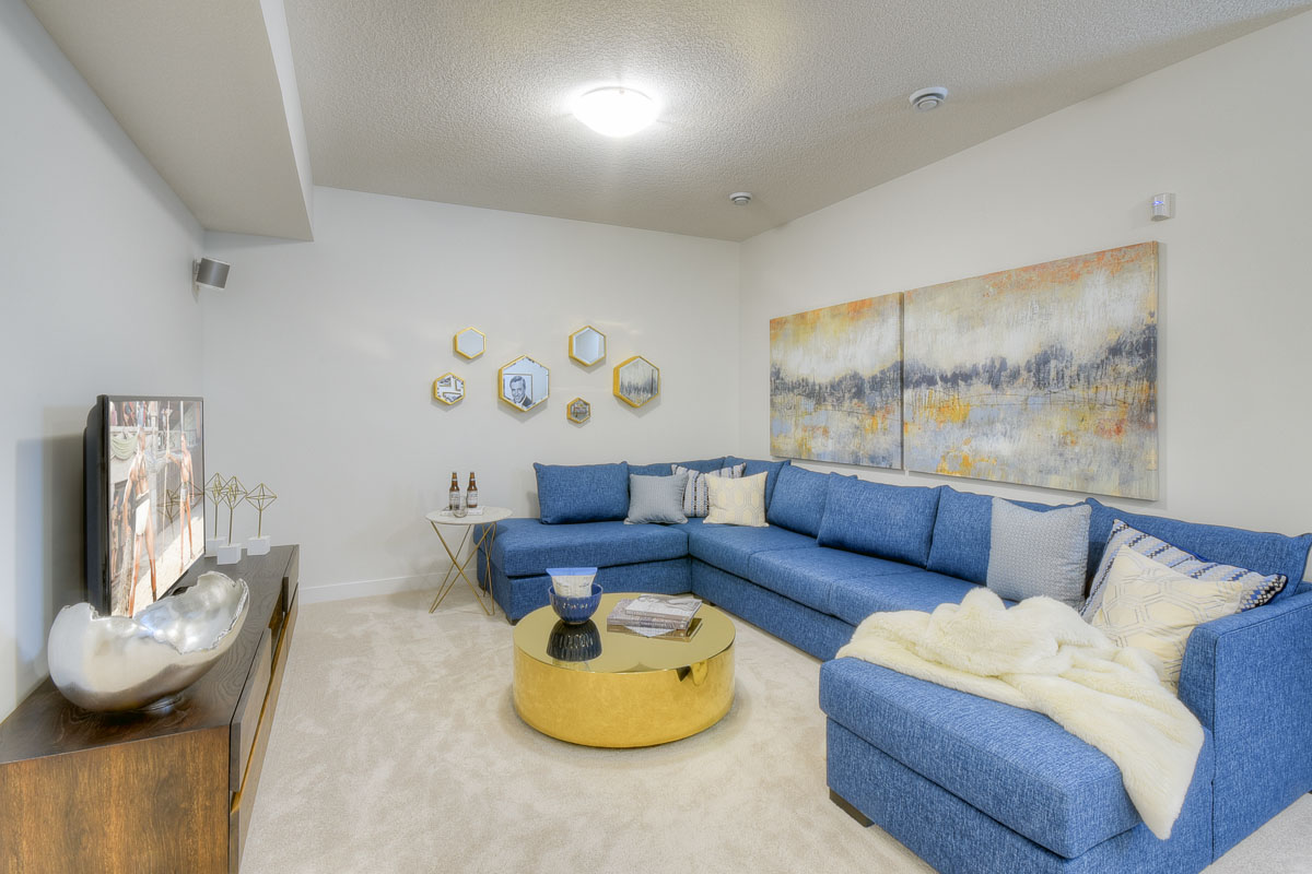 Basement living room with large blue sectional and round gold coloured coffee table in the Kingsley Cre model home.