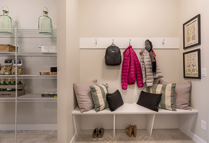 Mudroom next to pantry in the Kingston model home with a white bench and green and grey throw pillows.