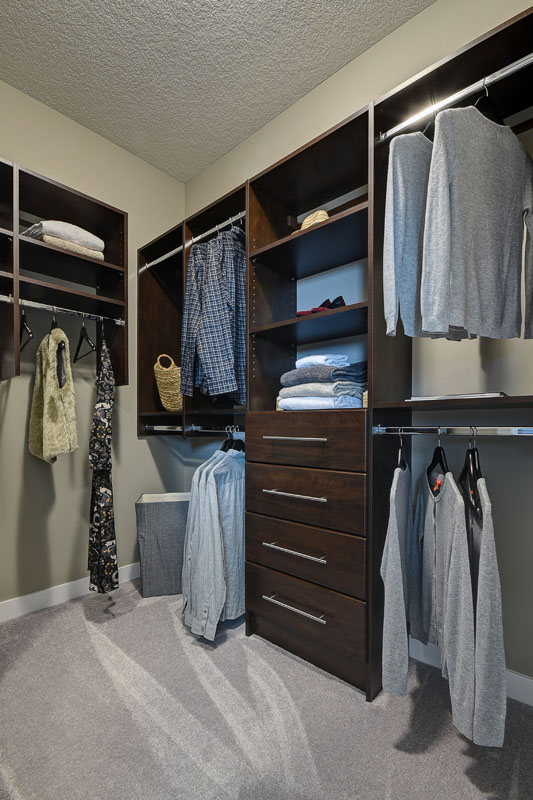 Walk in closet in the Saffron model home with tall brown dresser and built in shelving with clothes hanging.